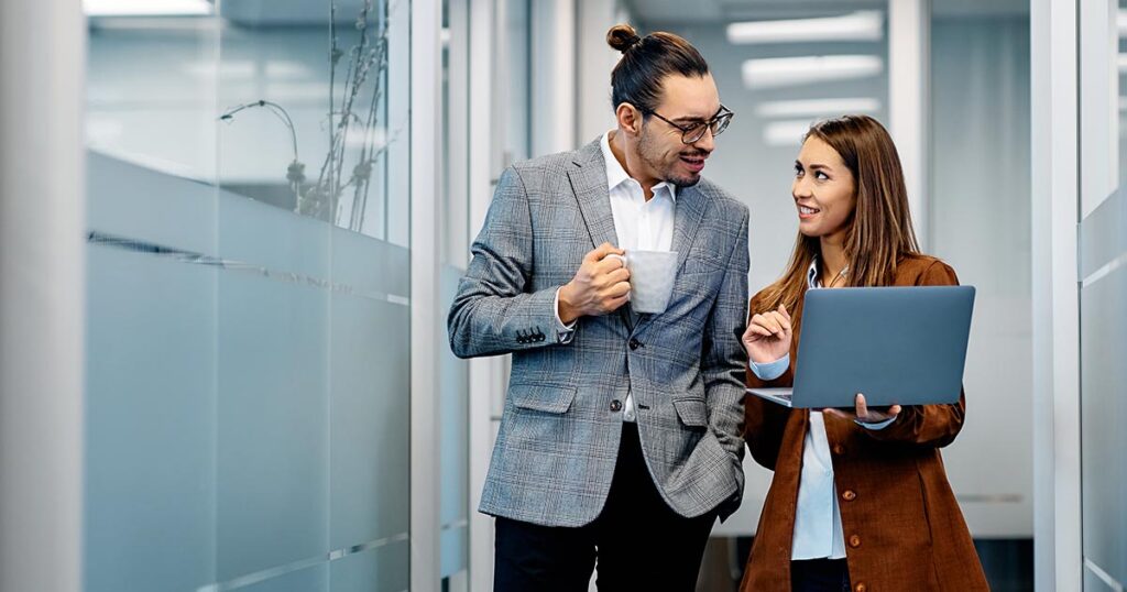 Two employees walk down the hall in an office setting looking at laptop.