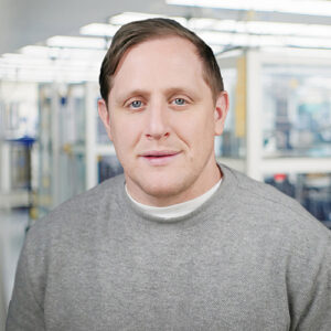 Chris Nikodem, BMS employee and engineer, stands outside a lab setting. He wears a grey top.